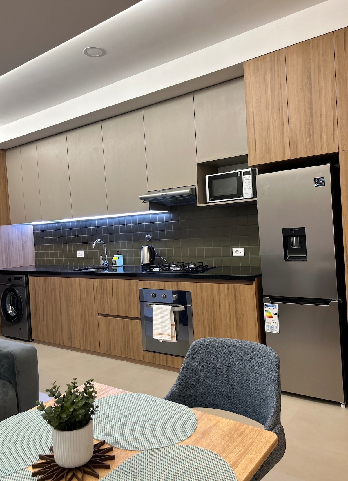 A modern kitchen area features sleek wooden cabinetry and energy-efficient appliances. A stainless steel refrigerator and microwave are positioned smoothly along the wall. The countertop is illuminated by under-cabinet lighting, enhancing the functional space that is complemented by a small dining table nearby.
