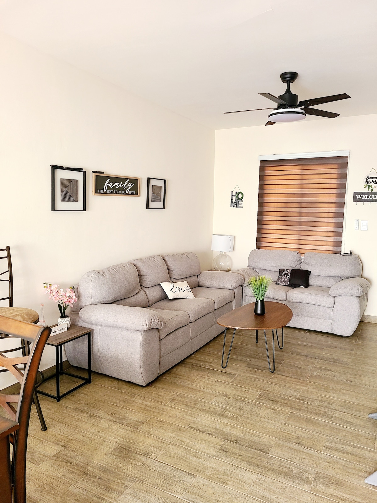 A living area features comfortable beige sofas arranged around a small circular table. Decorative wall frames and a potted plant add a touch of greenery. Light from a ceiling fan illuminates the space, complemented by natural light filtering through the window with blinds.