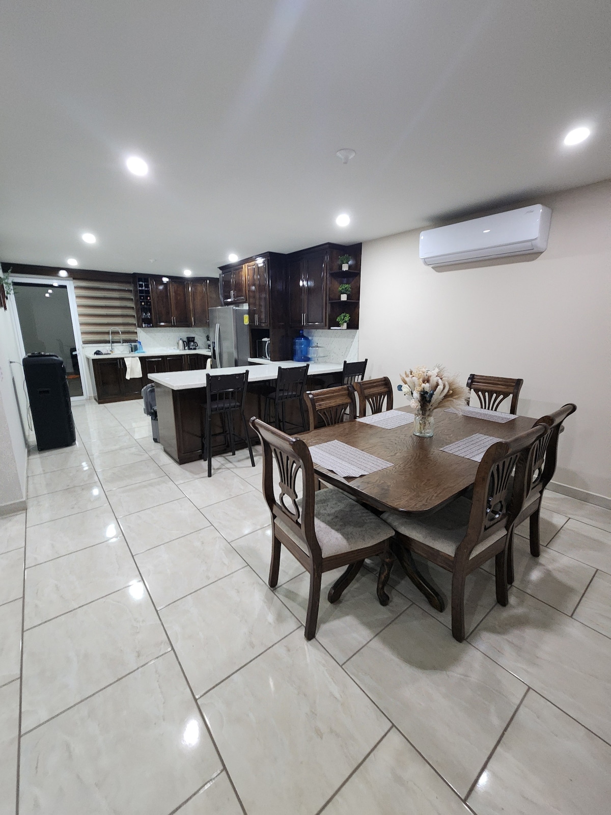The image features a modern kitchen and dining area with tile flooring. A wooden dining table with six chairs is situated in the foreground, and the kitchen is equipped with dark cabinets, a large island, and various appliances. Soft lighting is provided by ceiling fixtures.