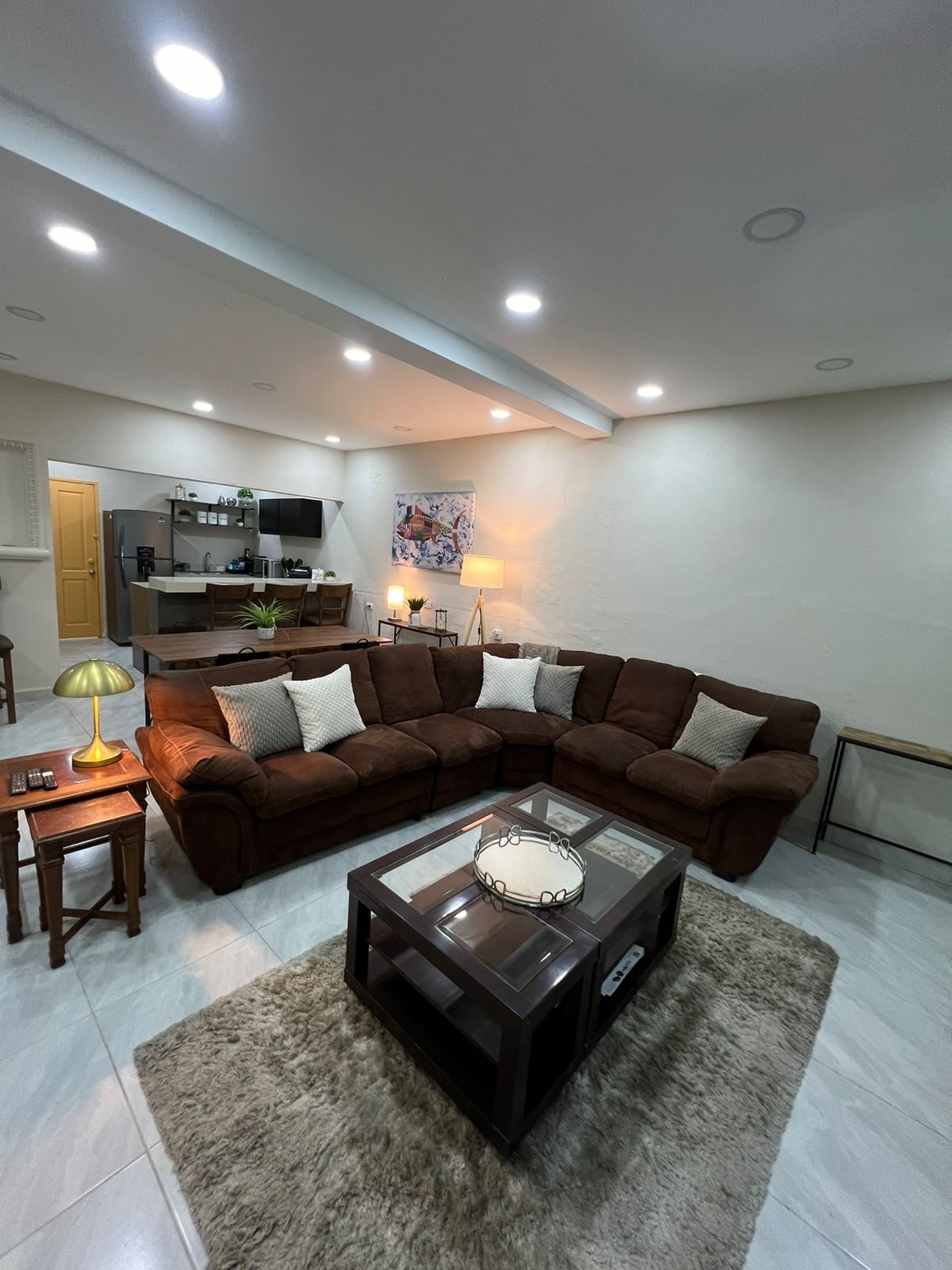 A spacious living area features a large, dark brown sectional sofa with several decorative cushions. A glass coffee table sits atop a soft area rug, while a lamp casts gentle light. The open kitchen is visible in the background, enhancing the communal feel of the space.