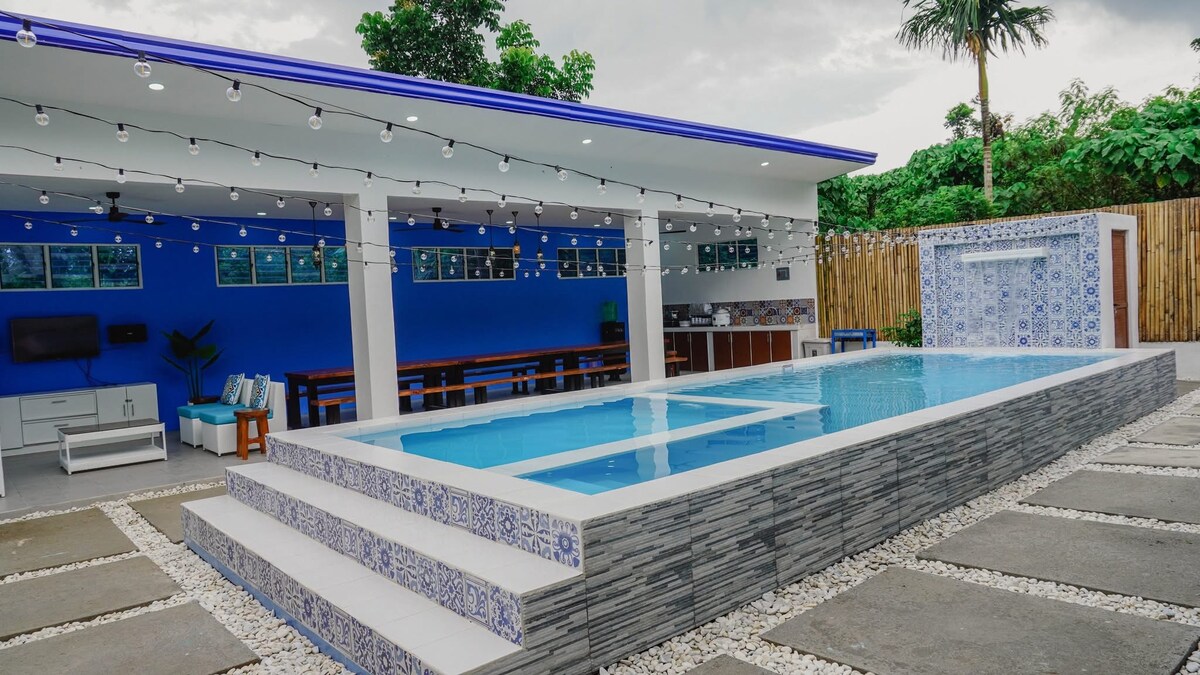 A spacious outdoor area features a modern swimming pool framed by decorative tiles. String lights are overhead, adding a subtle glow to the setting. The space includes a dining area with wooden tables and benches, alongside a kitchen area partially visible in the background.