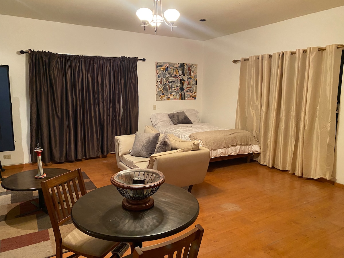 A spacious living area is furnished with a seating arrangement that includes a cozy chair and a dining table set for four. A comfortable bed is positioned against the wall, with soft curtains framing the windows. Artwork adds character to the room.