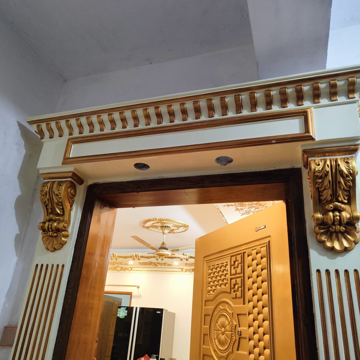 An ornate doorframe features intricate gold detailing and decorative carvings. The door itself is a rich gold color with a textured design. A glimpse of a bright room is visible through the doorway, showcasing a modern kitchen space with appliances in the background.