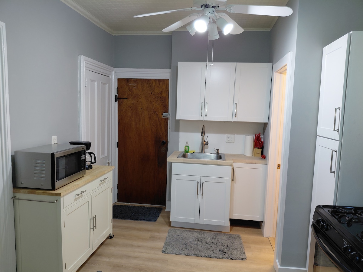 A well-equipped kitchen features white cabinetry, a stainless steel sink, and a countertop with essential appliances. A microwave and a wooden serving table are positioned nearby, while a ceiling fan provides ventilation. Soft lighting enhances the welcoming atmosphere.