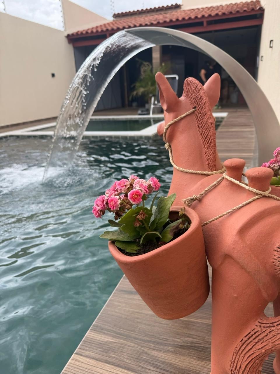 A decorative clay horse stands beside a pool, holding a planter filled with pink flowers. The pool features an elegant water feature, with water arching gracefully into the water below. A sunlit patio area is visible in the background, enhancing the tranquil setting.