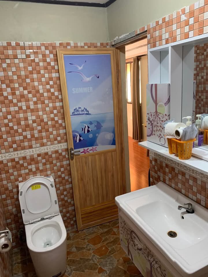 A clean bathroom features tiled walls in warm tones. A wooden door with an ocean-themed design leads to another area. A white sink is set against a matching countertop, and a toilet is positioned nearby. Natural light filters through a window, illuminating the space.