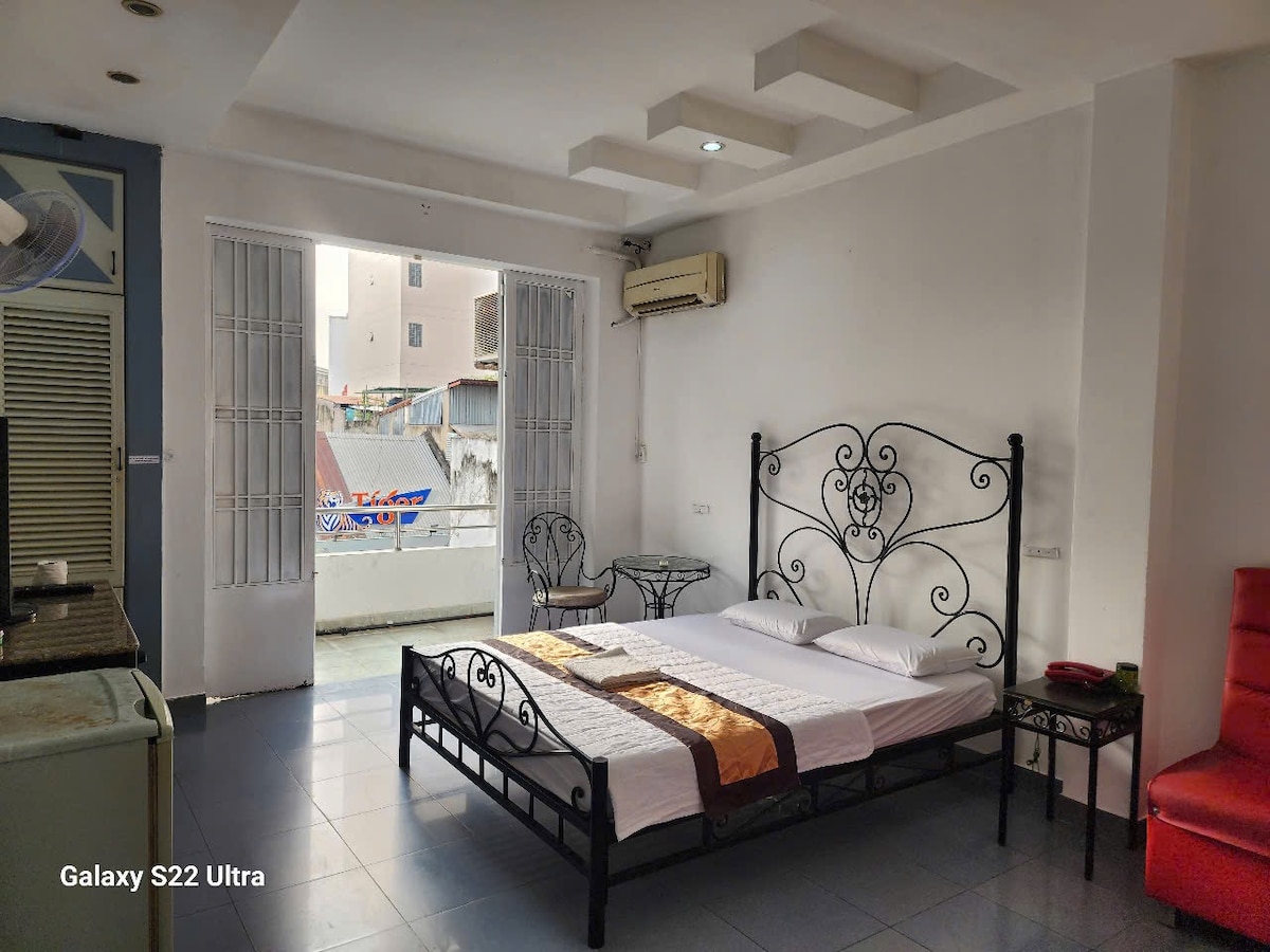 A spacious room features a black wrought iron bed with a patterned bedspread. Light enters through two large windows with sheer curtains, illuminating the tiled floor. A small balcony is visible, along with a kitchenette area in the background.