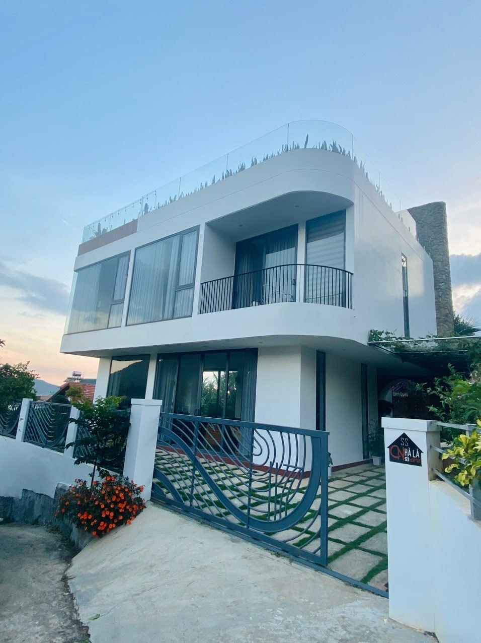 A modern villa is showcased with a sleek exterior design featuring large glass windows and a spacious balcony. Lush greenery and colorful flowers enhance the entrance, while a well-maintained pathway leads up to the home, framed by a stylish metal fence.