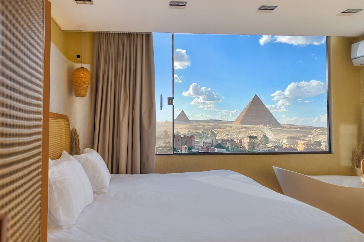 A spacious bed is visible, positioned near a large window that frames the iconic view of the Giza Pyramids. Soft, neutral-toned décor complements the natural light flooding the room, while gentle patterns of fabric add texture to the space.