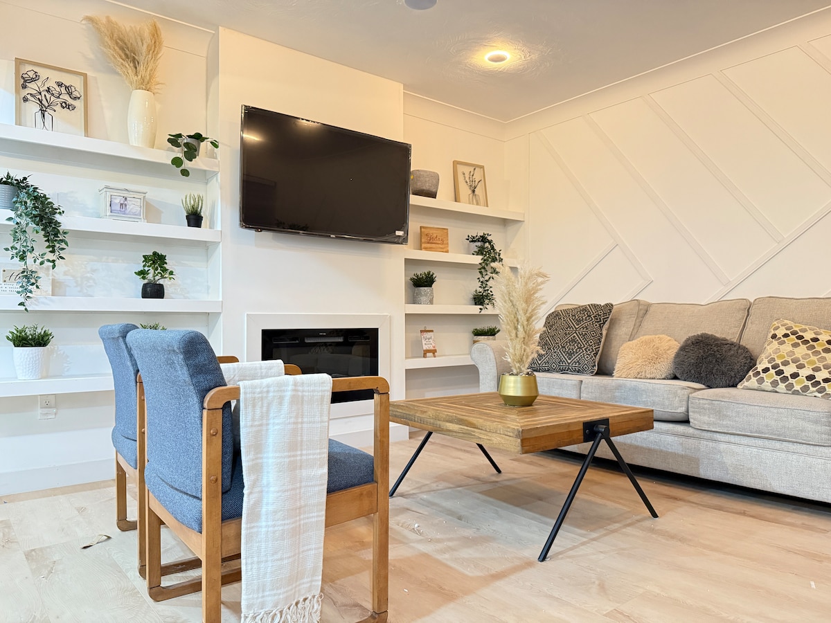 A light and airy living area is featured, showcasing a modern wooden coffee table at the center. A comfortable sectional sofa is adorned with textured cushions. Shelves with decorative plants frame the space, while a wall-mounted television and a minimalist fireplace add to the cozy ambiance.
