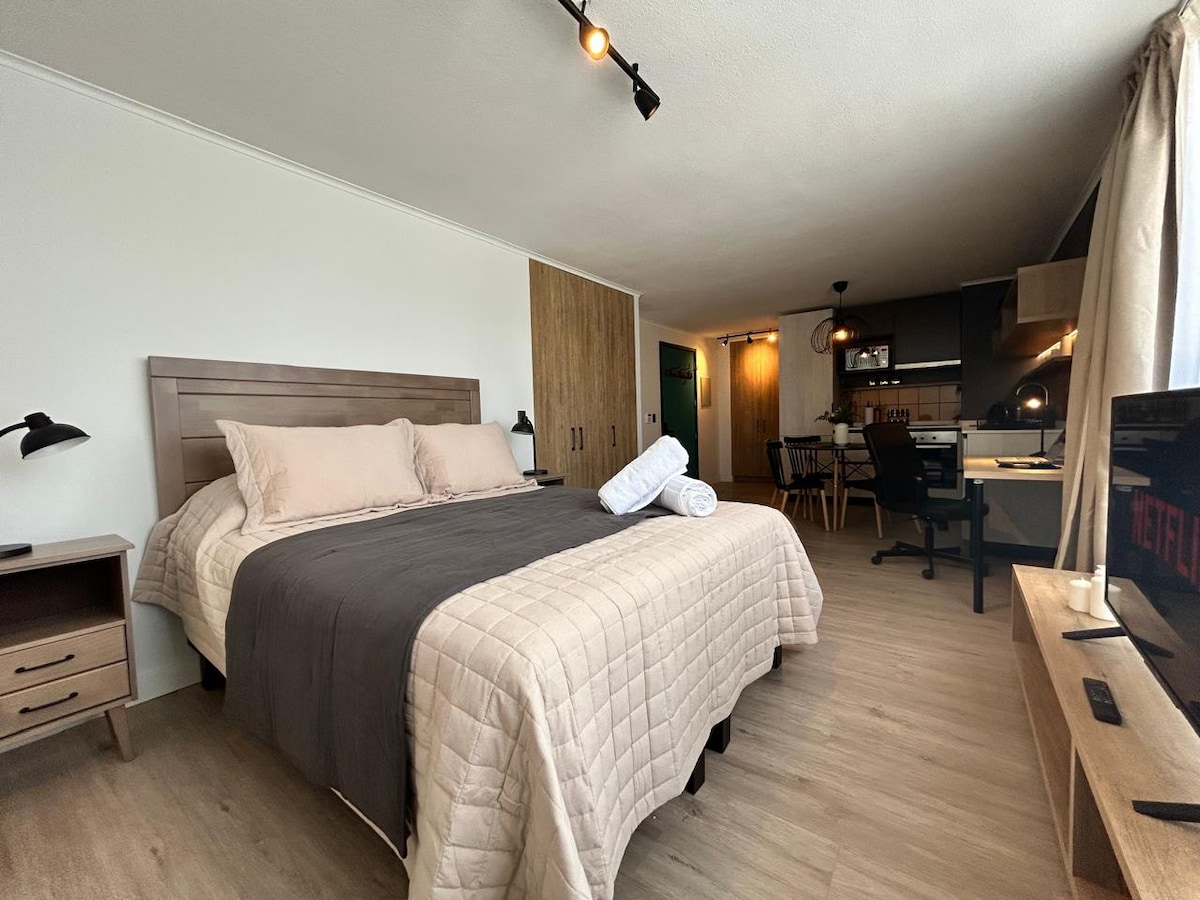 A spacious sleeping area is highlighted by a large bed adorned with neutral-colored linens. Two pillows lie at the head of the bed, alongside a bedside table with a lamp. The room features a light wood floor, a kitchenette in the background, and a television on a wooden console.