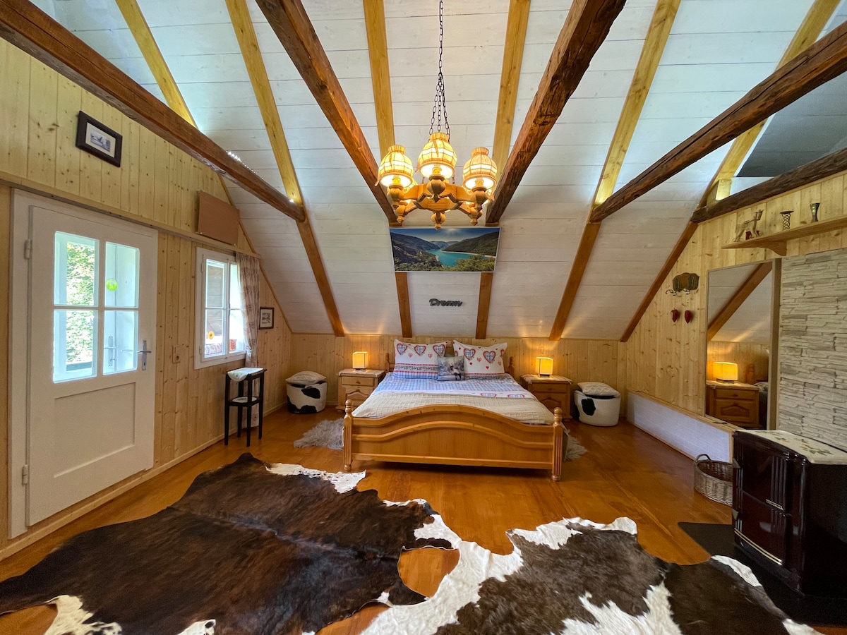 A spacious bedroom features wooden beams across the ceiling and natural wood walls. A large bed is centered in the room, flanked by bedside lamps. A cowhide rug is placed on the wooden floor, while windows provide gentle natural light.