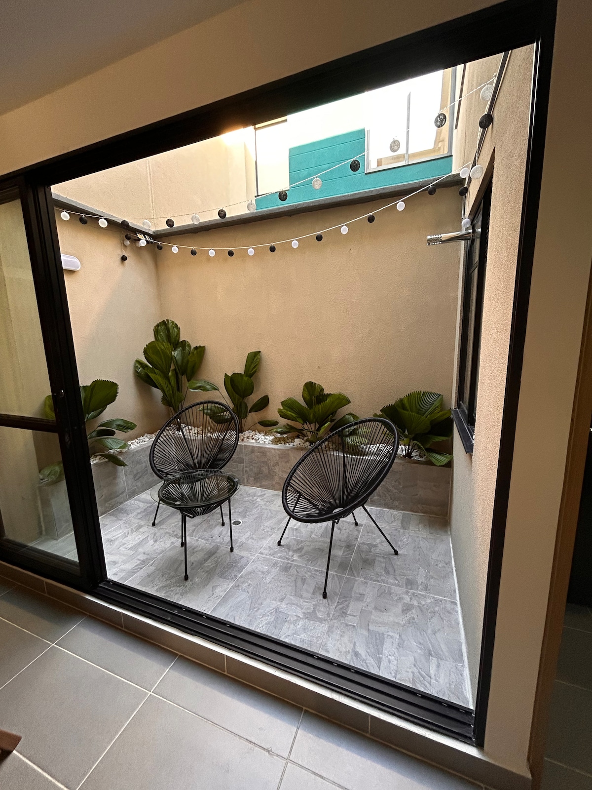 A small outdoor seating area is presented, featuring two black wire chairs positioned on a tiled floor. Potted plants border the space, complemented by string lights overhead, creating a cozy atmosphere. Natural light enters through the glass doors, enhancing the inviting environment.