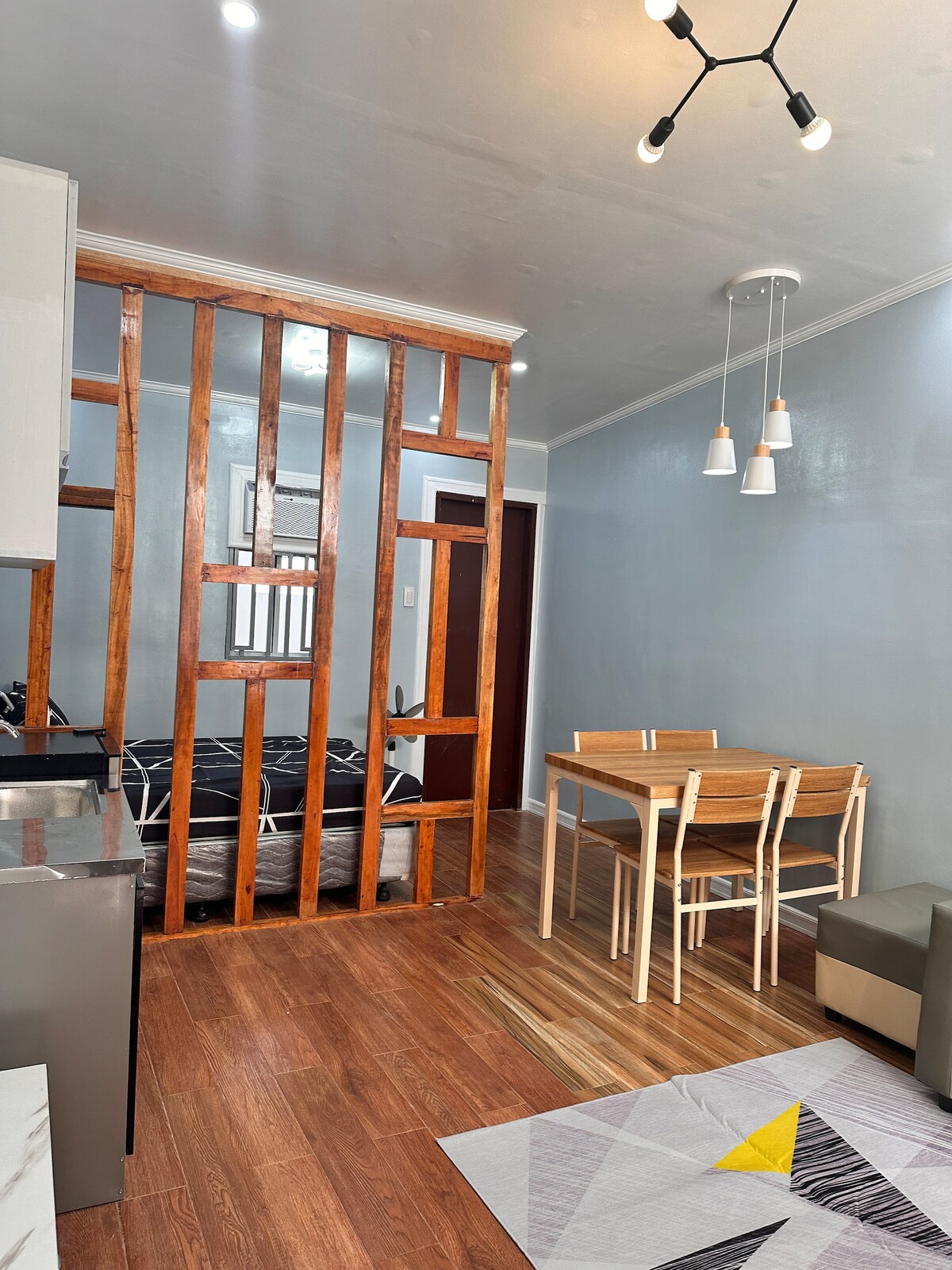 An open living area is divided by wooden slats, showcasing a sleek kitchen on one side and a cozy sleeping space with a patterned bed cover on the other. A dining table with four chairs sits adjacent to the sofa, with a modern light fixture overhead.
