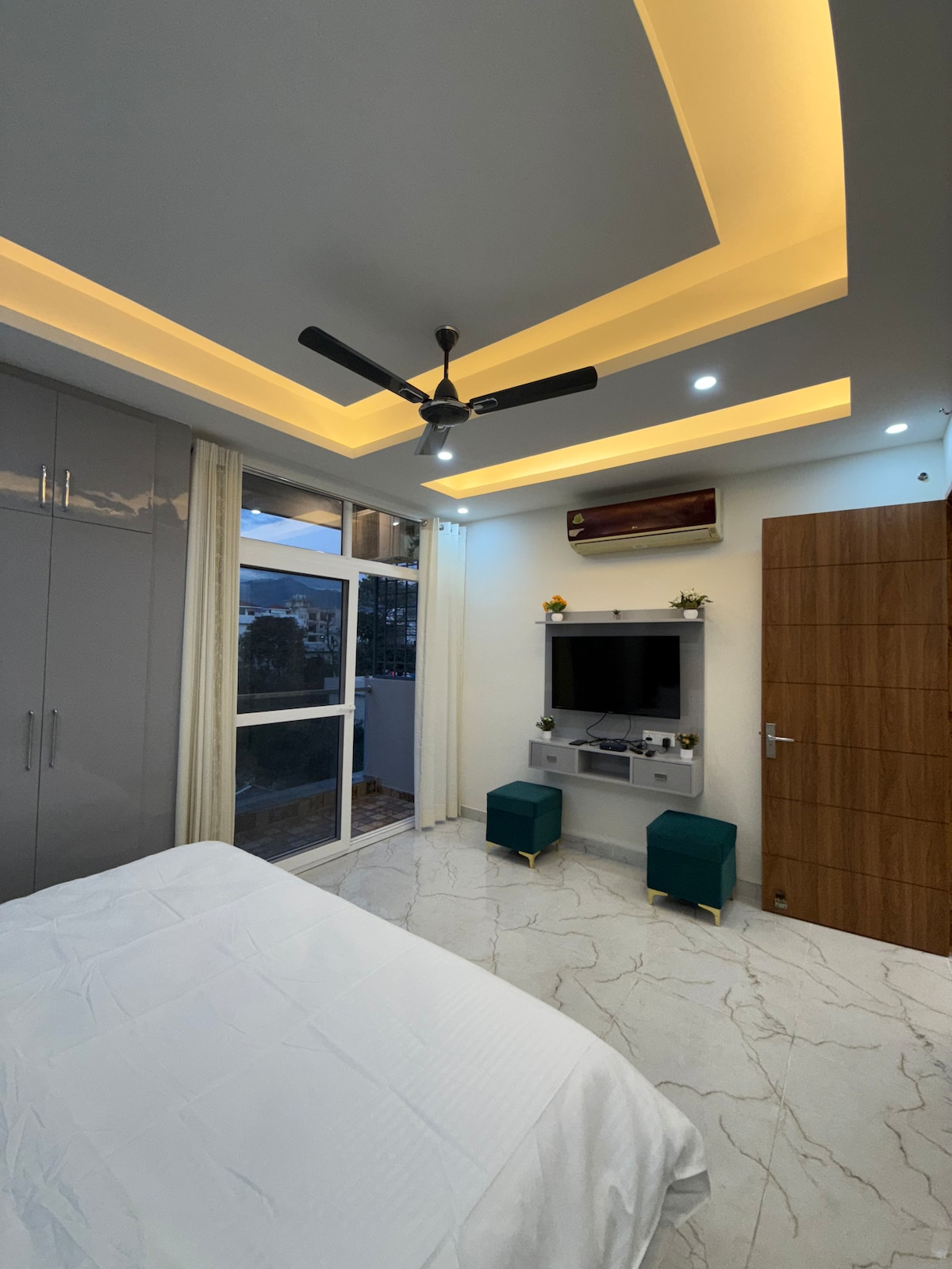 A serene bedroom featuring a spacious bed with light bedding. A ceiling fan and recessed lighting enhance the ambiance. A wall-mounted television sits above a console table, flanked by two teal stools. Large glass doors lead to an outdoor view, with soft curtains adding a touch of privacy.