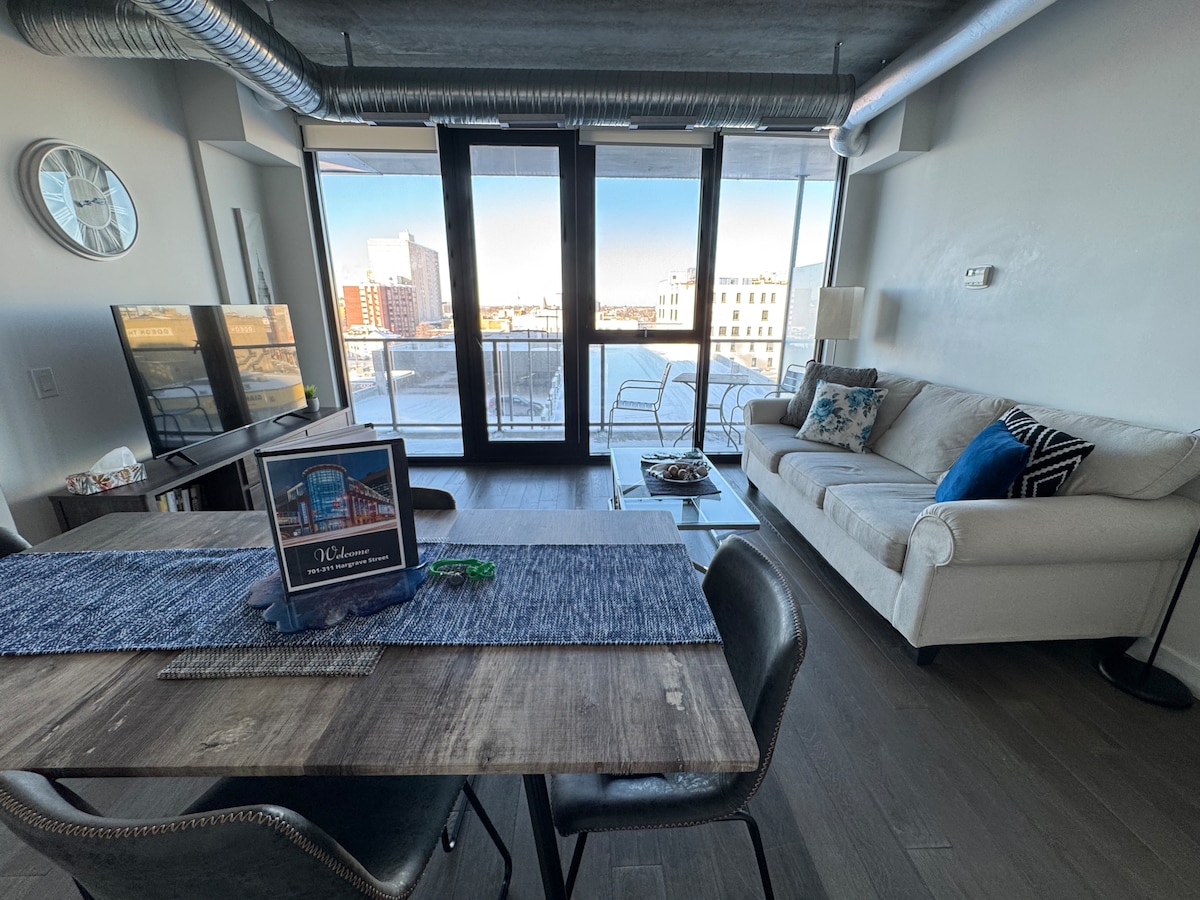 A bright living space features a spacious couch accented with decorative pillows and a large window offering outdoor views. A dining table with modern chairs is positioned nearby, topped with a blue runner. A television and coffee table complete the inviting atmosphere.