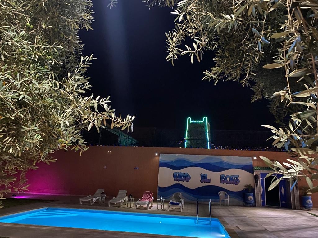 A swimming pool is framed by lush olive trees, with soft lighting creating a serene atmosphere. In the background, subtle illumination outlines the structure, adding visual interest to the scene at night. Lounge chairs are positioned nearby, inviting relaxation.