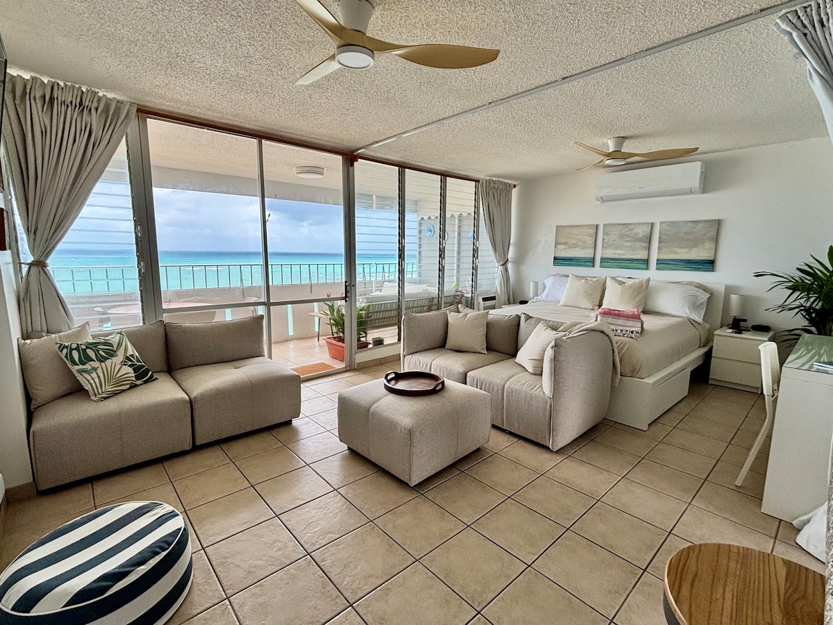 The studio features a light-filled open layout with a king-sized bed and a cozy seating area. Soft, neutral-toned furnishings are complemented by plant decor. Large windows provide expansive views of Playa Azul, with sheer curtains allowing soft natural light to gracefully illuminate the space.