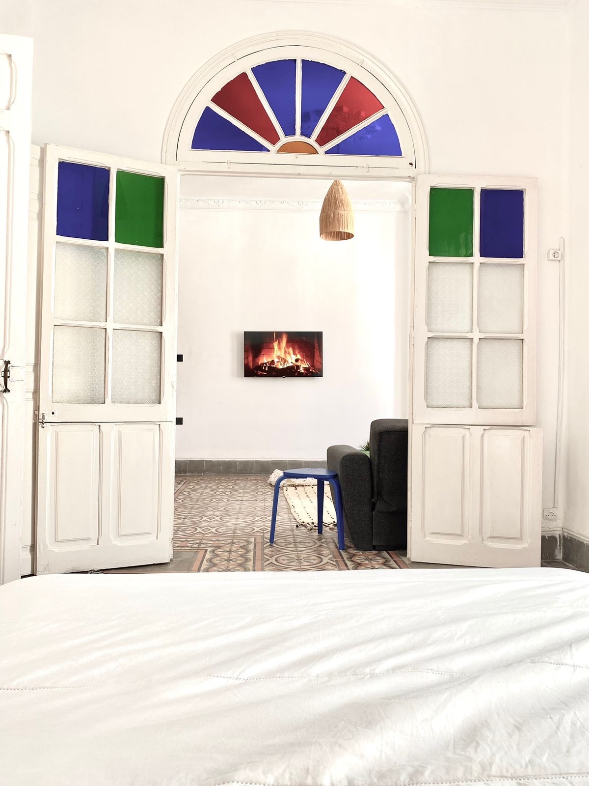 A room is entered through double wooden doors featuring colorful glass panels. A cozy seating area is visible beyond, with a small blue table and a dark sofa. A wall-mounted electric fireplace adds warmth to the space, complemented by patterned tile flooring.