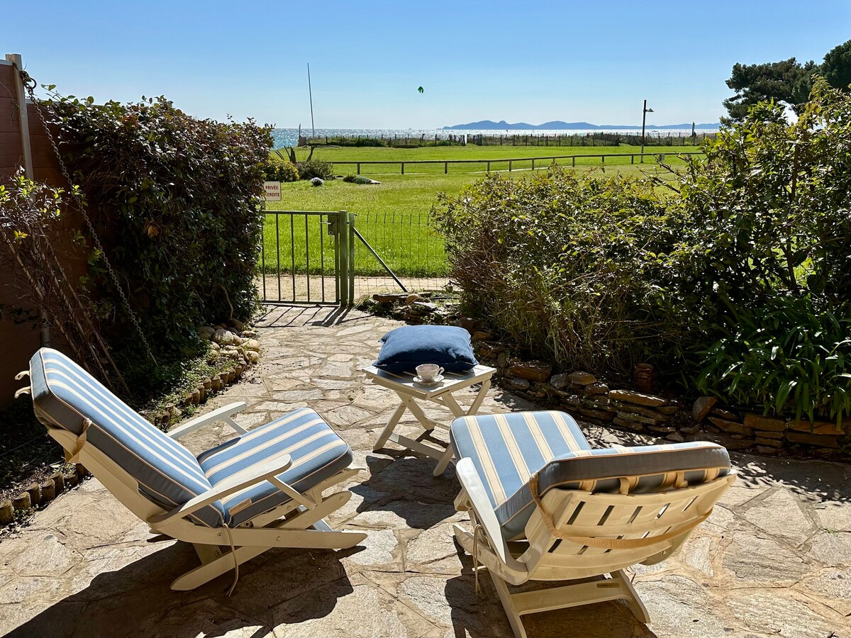 A serene patio area features two striped lounge chairs facing a grassy expanse, leading to an unobstructed view of the sea. A small table rests between the chairs, adding a space for relaxation. Lush greenery surrounds the area, enhancing the outdoor setting.