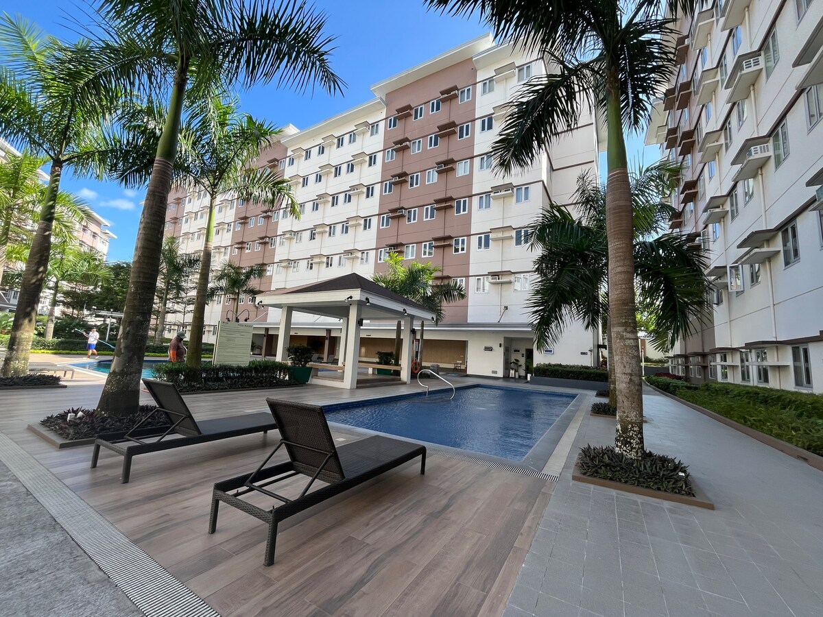 An inviting pool area is bordered by tall palm trees and neatly arranged lounge chairs. A shaded pavilion is visible nearby, enhancing the communal space. Surrounding the pool, well-maintained landscaping adds to the area's tranquility, with a modern multi-story building in the background.