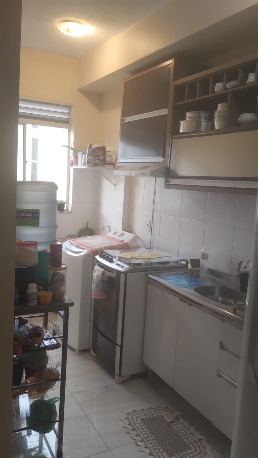 A compact kitchen is equipped with essential appliances, including a washer and a stove. Cabinets provide storage space, while shelves display dishware. Natural light enters through a nearby window, enhancing a bright and functional cooking area.