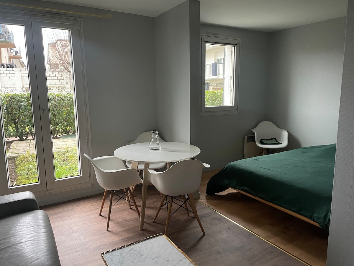 A bright living space features a cozy sleeping area with a green bedspread and a small dining table with chairs. Large windows allow natural light and provide a view of the garden outside, enhancing the room's connection to nature.