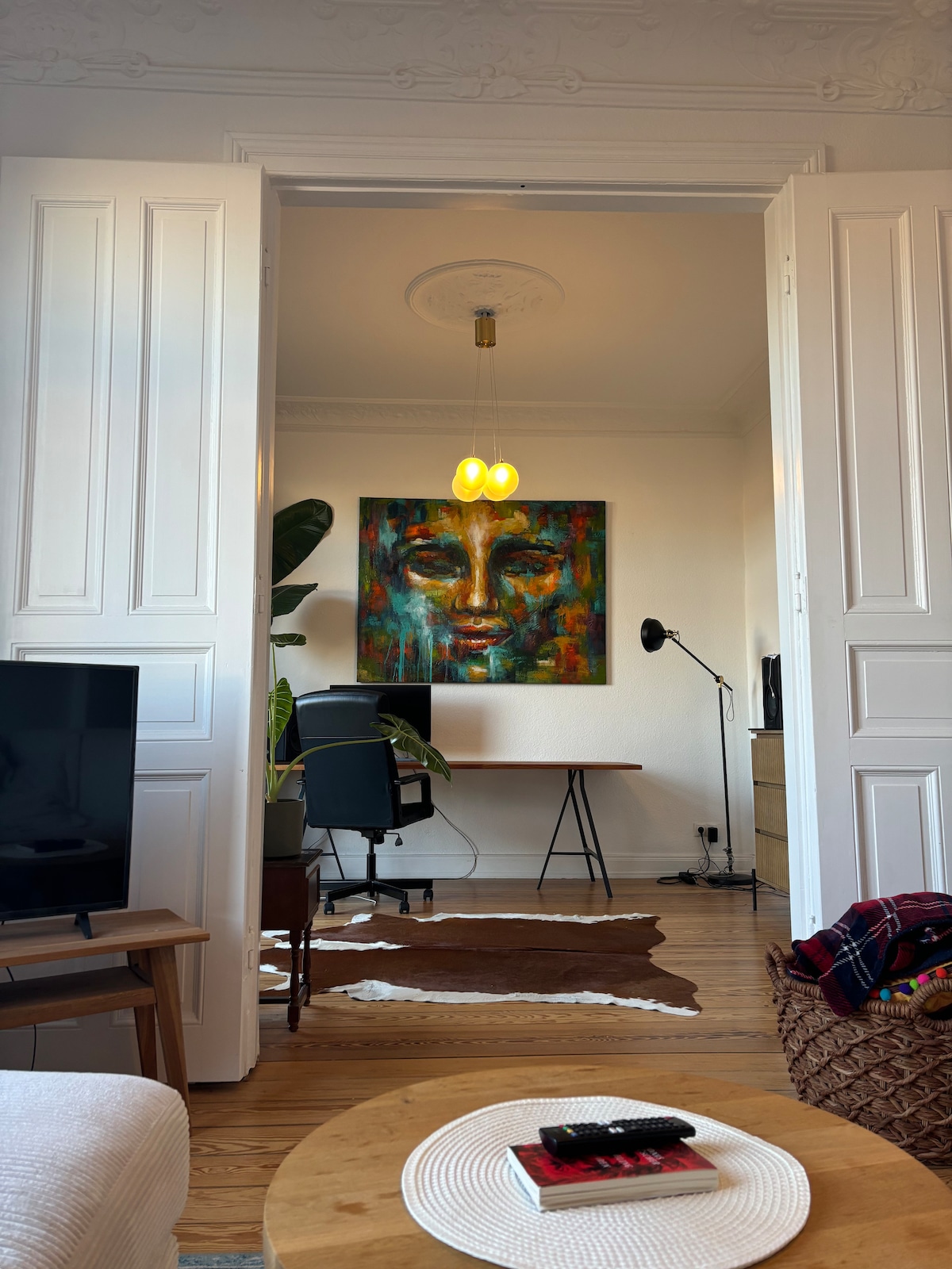 An open doorway frames a light-filled workspace featuring a desk and a chair. A decorative wall art adds color to the background, while a floor lamp provides illumination. The foreground includes a cozy living area with a low table and a soft rug.