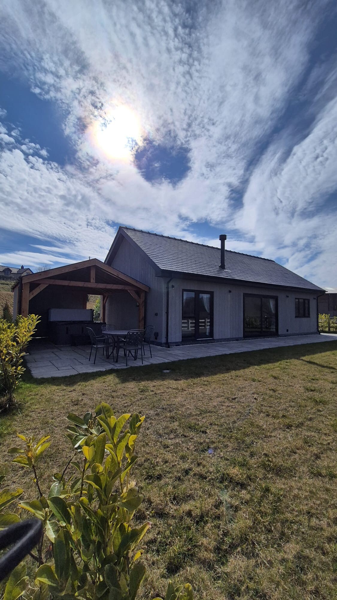 The exterior of the lodge is shown, featuring a covered outdoor area with seating and a hot tub. A spacious lawn surrounds the property, and an expansive sky fills the background with scattered clouds, providing a serene and open atmosphere.