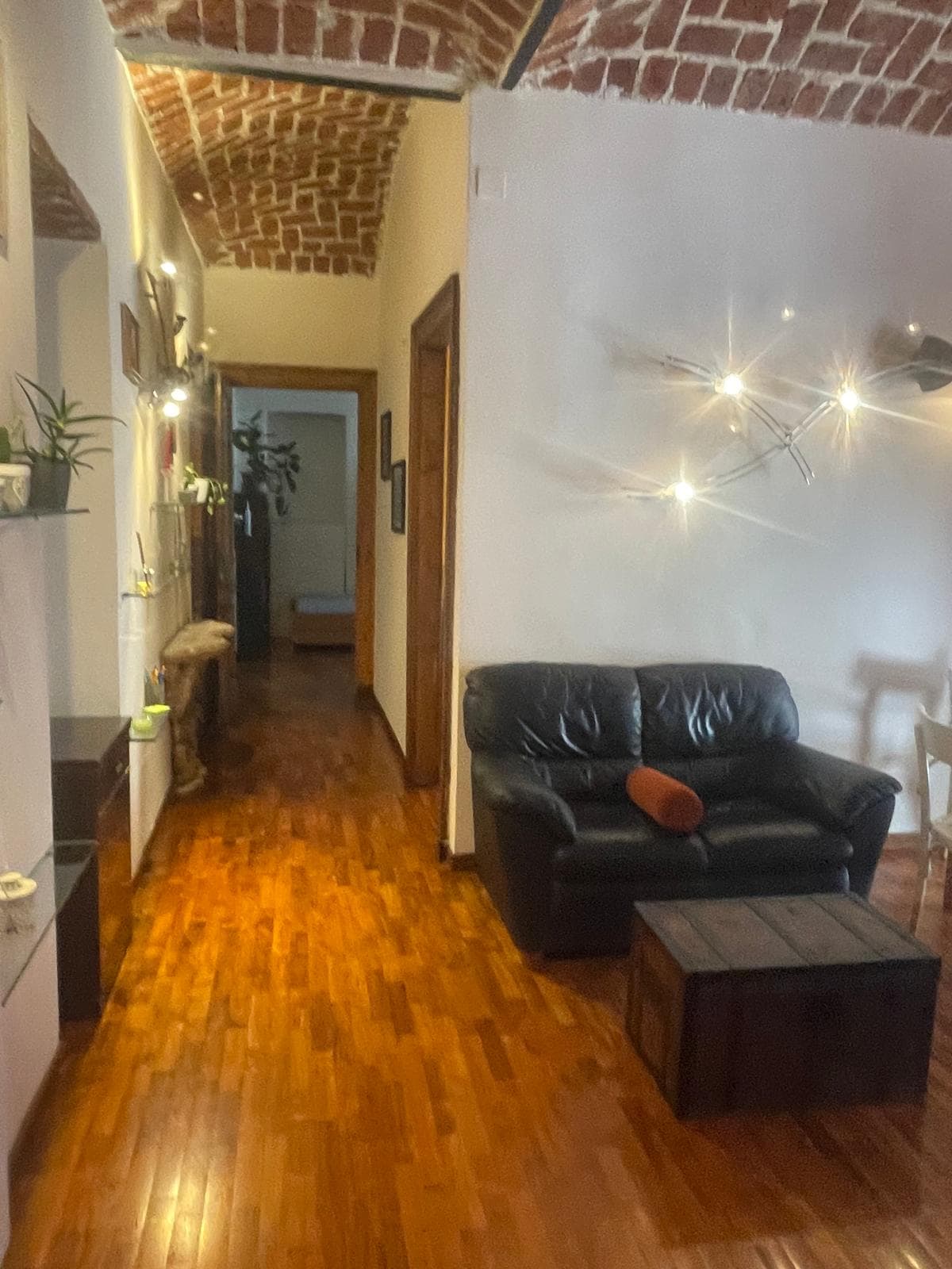 A narrow hallway features warm wooden flooring, leading to various rooms. A black leather sofa is positioned beside a wooden coffee table, creating a cozy seating area. Soft lighting is provided by minimalist fixtures mounted on the wall, with decorative plants accentuating the space.