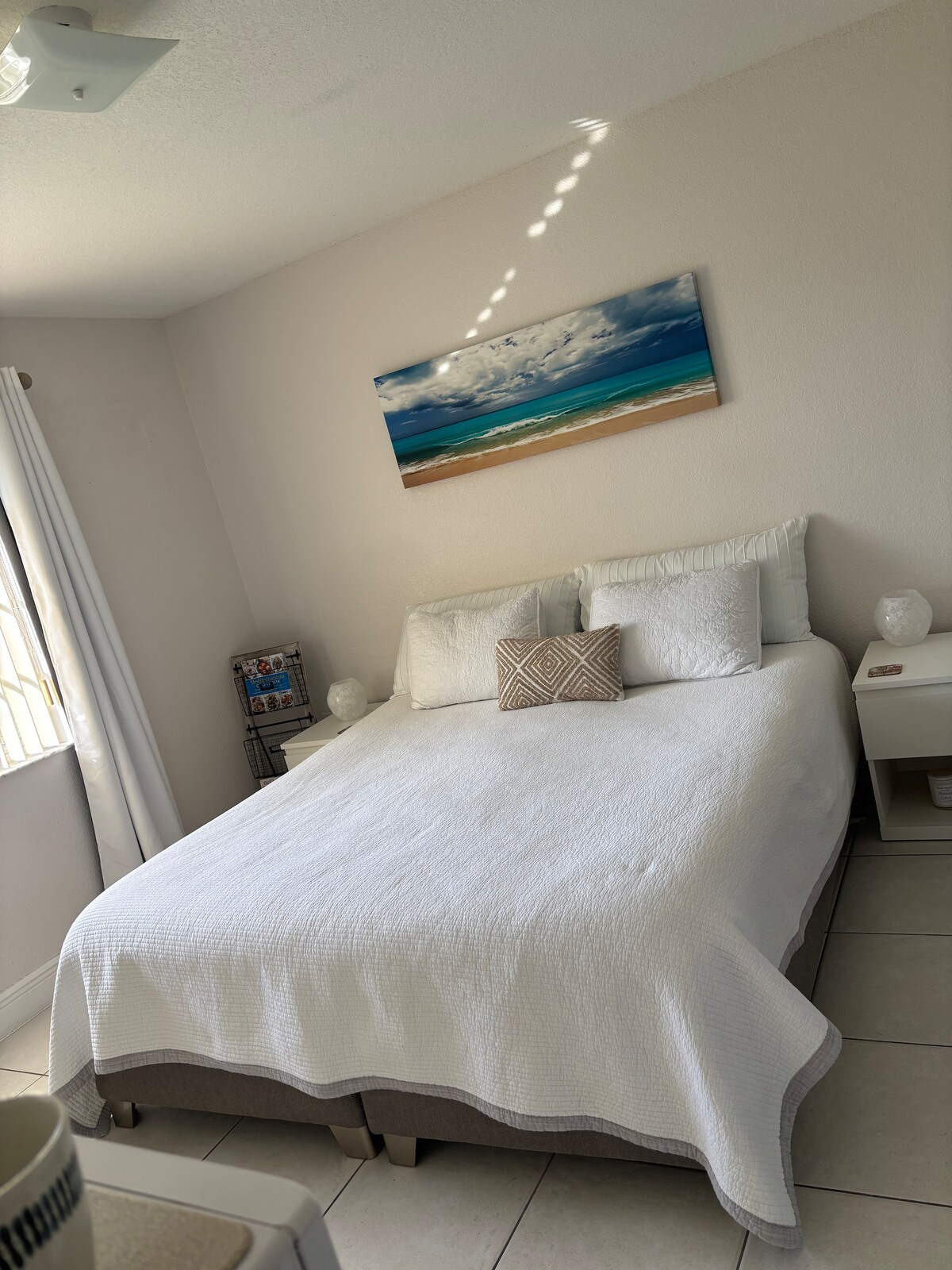 A bright sleeping area features a spacious bed dressed in a white quilt with decorative pillows. Light-colored walls and a piece of wall art depicting a beach scene enhance the calming environment. Nightstands are positioned on either side of the bed, each holding a lamp.