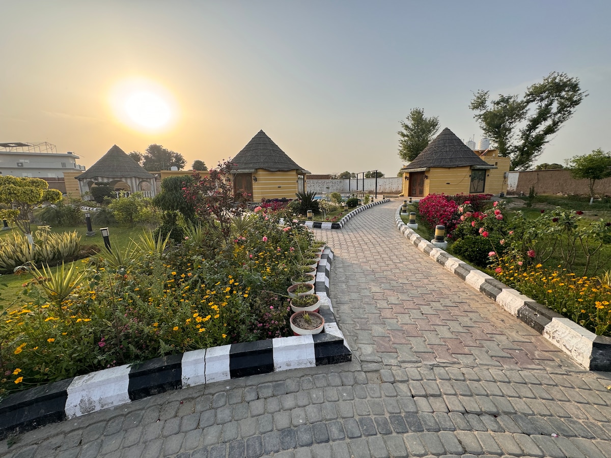 A path is bordered by vibrant flowers and greenery, leading to two charming cottages with conical roofs. The sun sets in the background, casting a warm glow over the serene landscape, while the cobblestone walkway guides visitors through the picturesque garden.