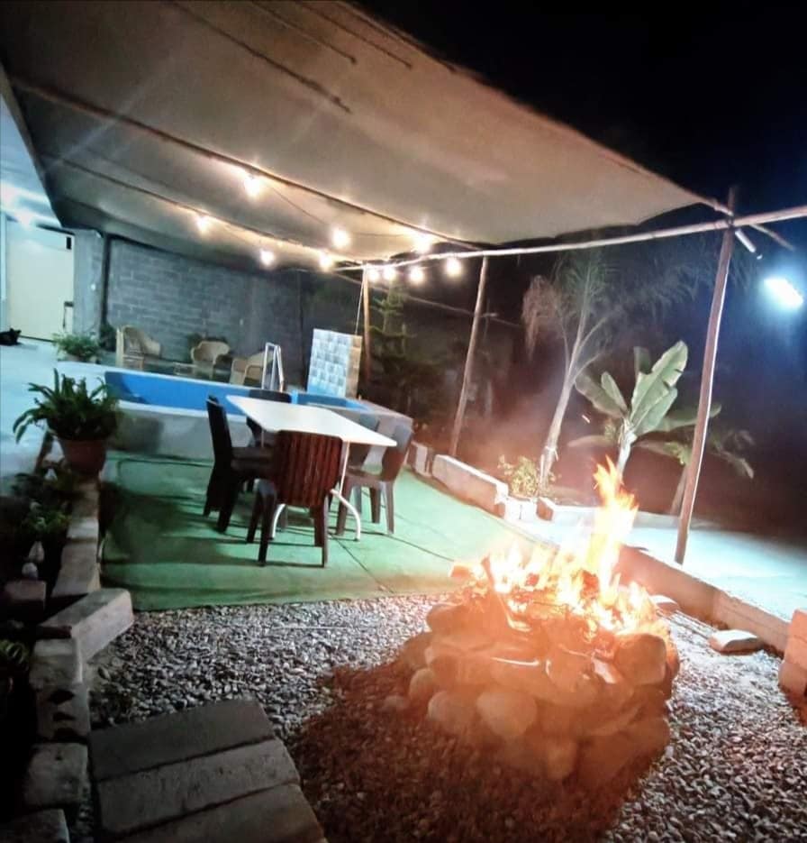 An outdoor area is shown, featuring a stone fire pit surrounded by rocks. A table with chairs is placed nearby, and a pool is visible in the background. String lights illuminate the space, creating a cozy ambiance among surrounding greenery.