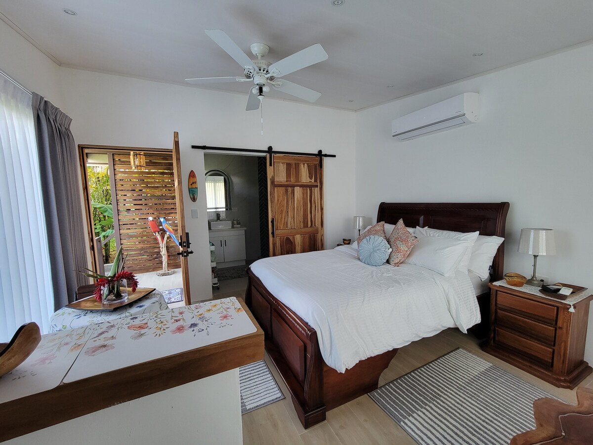 A spacious bedroom features a queen-size bed covered with crisp white linens and decorative pillows. Light wood accents are paired with a ceiling fan, while a large window offers natural light. A sliding door leads to a private outdoor patio, enhancing the sense of openness.