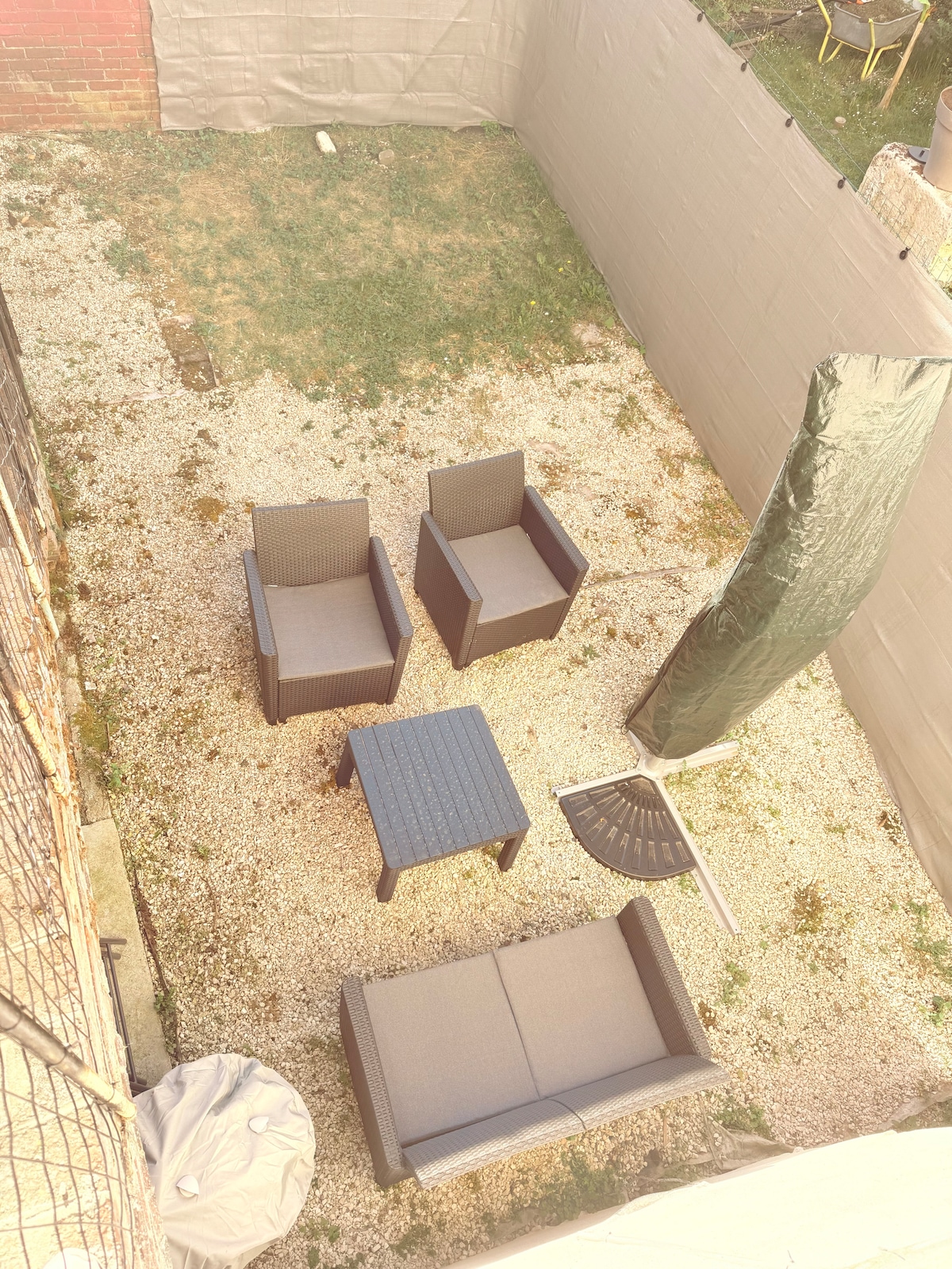 An outdoor courtyard is visible, featuring a seating arrangement with two armchairs and a sofa surrounding a center table. The area is paved with gravel and includes a protective cover for an unassembled umbrella. A portion of grass adds a touch of greenery to the space.
