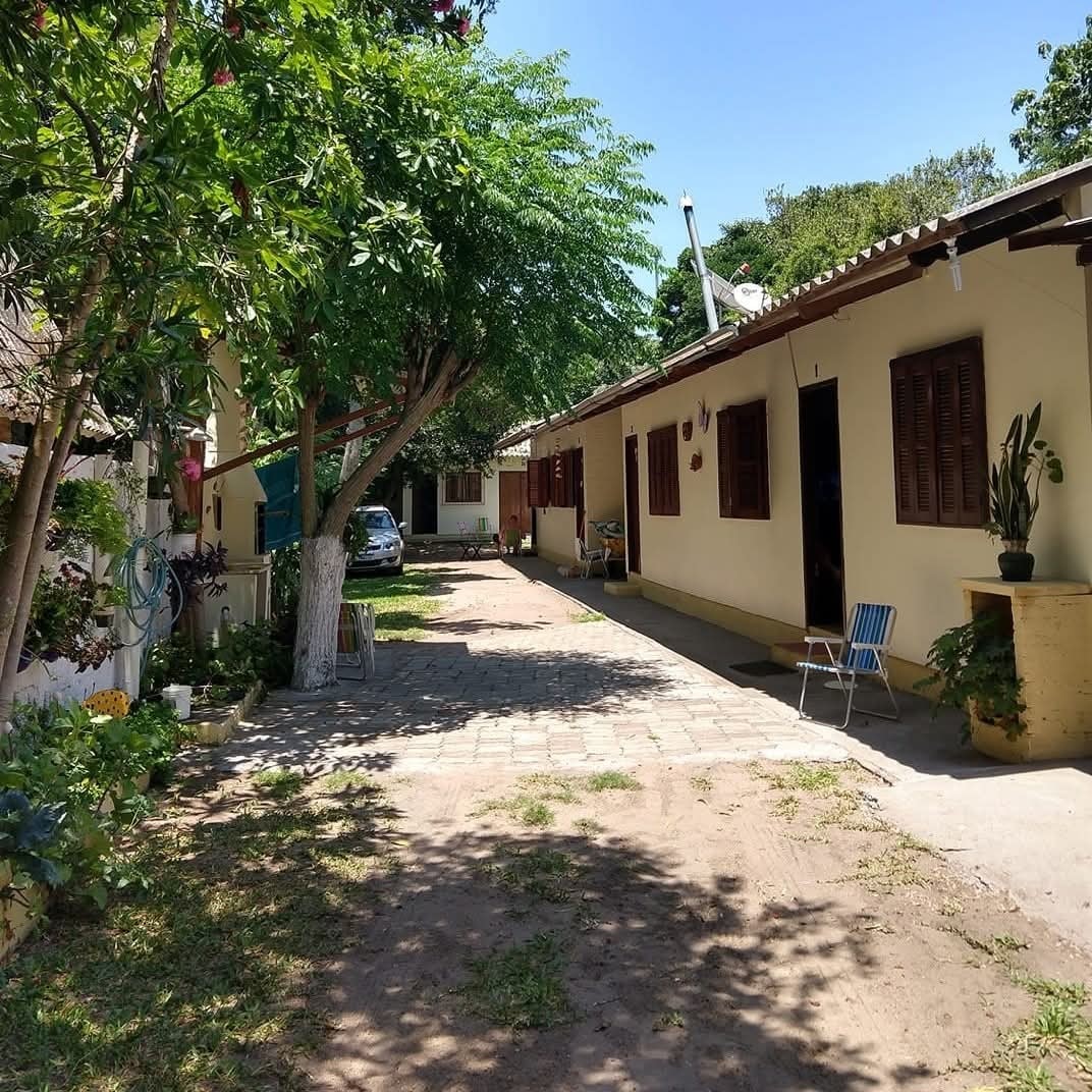 A shaded pathway is lined with manicured greenery and flowering plants, leading to two single-story buildings. The space includes a parked vehicle and a blue folding chair placed beside a plant-filled planter.