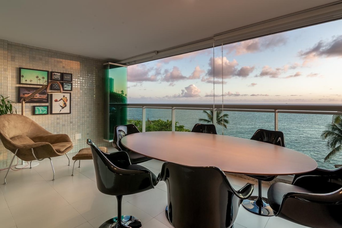 A dining area features an oval table surrounded by black armchairs, situated near expansive glass windows that offer an unobstructed view of the ocean. A cozy armchair is positioned in the corner, complementing the tranquil atmosphere created by the seascape visible from the interior.