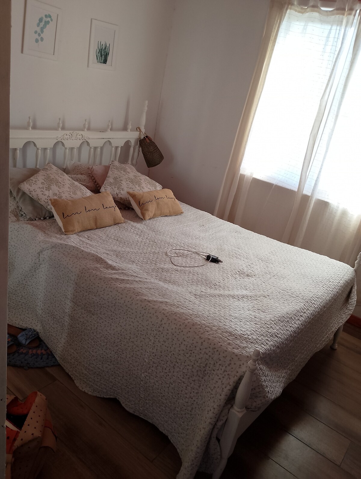 A cozy sleeping area features a white bed frame adorned with a light quilt. Two decorative pillows with text sit among neutral-toned bedding. Natural light filters through sheer curtains, illuminating the simple, serene décor, while wood flooring adds warmth to the space.