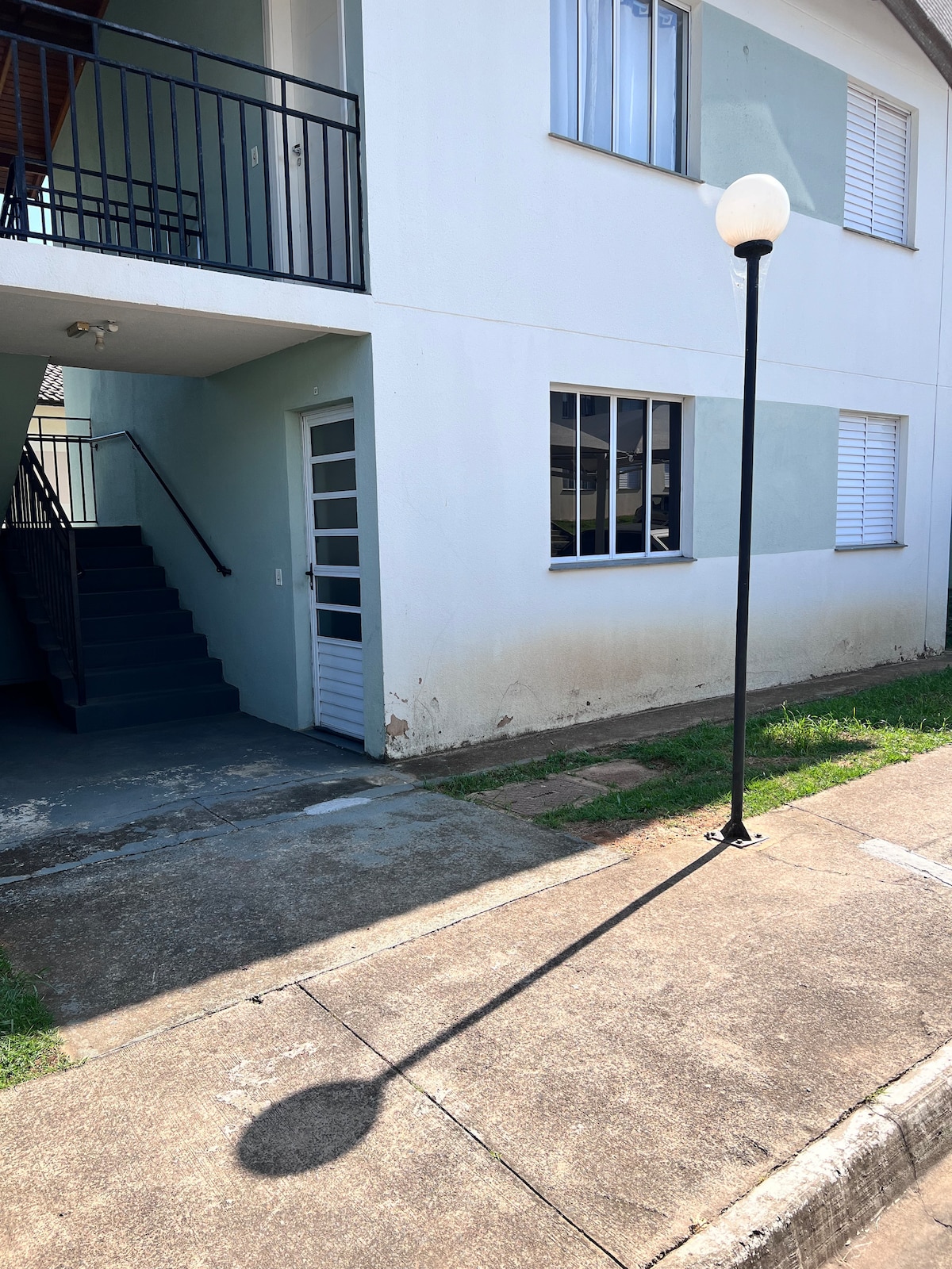 The exterior view of a two-story apartment building is shown, featuring a white facade and light blue accents. A set of stairs leads to the upper floor, while two windows are visible on the ground level. A lamppost stands beside a paved pathway.