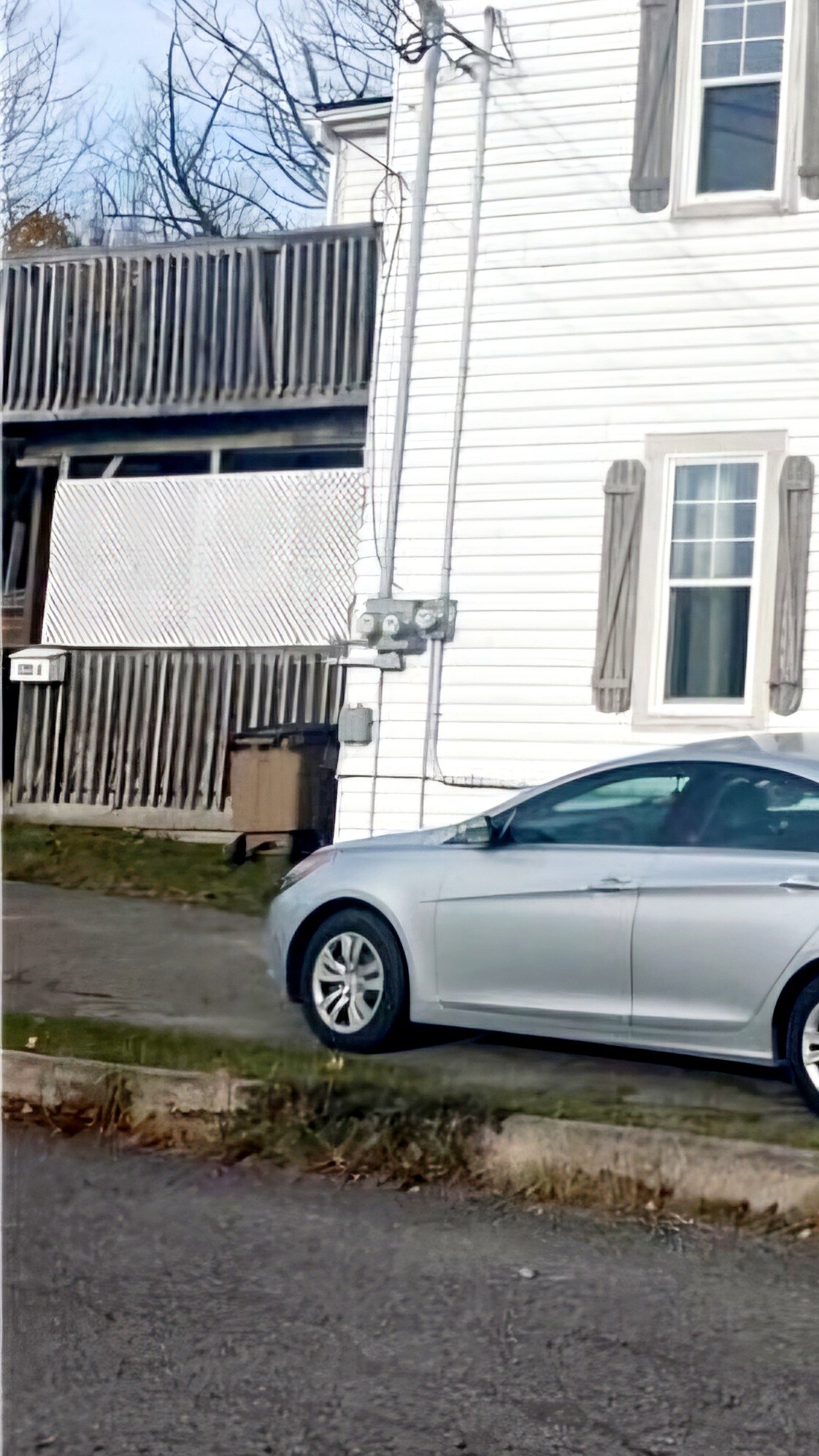 A silver car is parked along a tree-lined street beside a white house with distinct grey shutters. The building features a raised deck with a lattice design, and utility meters are visible on the lower wall. Natural light highlights the car and surroundings.