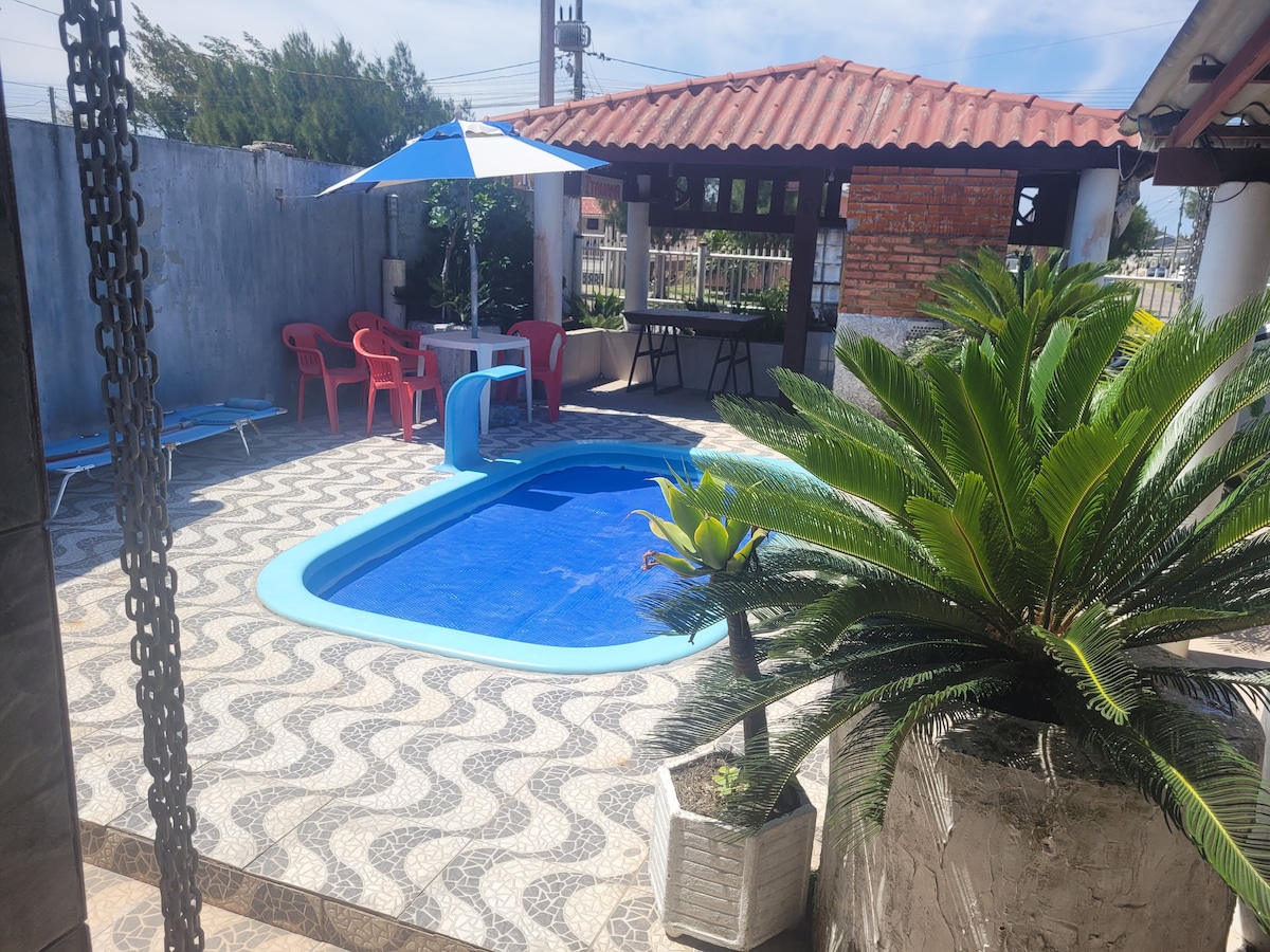 A vibrant outdoor area is showcased, featuring a blue pool surrounded by patterned tiles. Red chairs offer seating, complemented by an umbrella for shade. Lush greenery is present in planters, adding to the inviting atmosphere of the space.