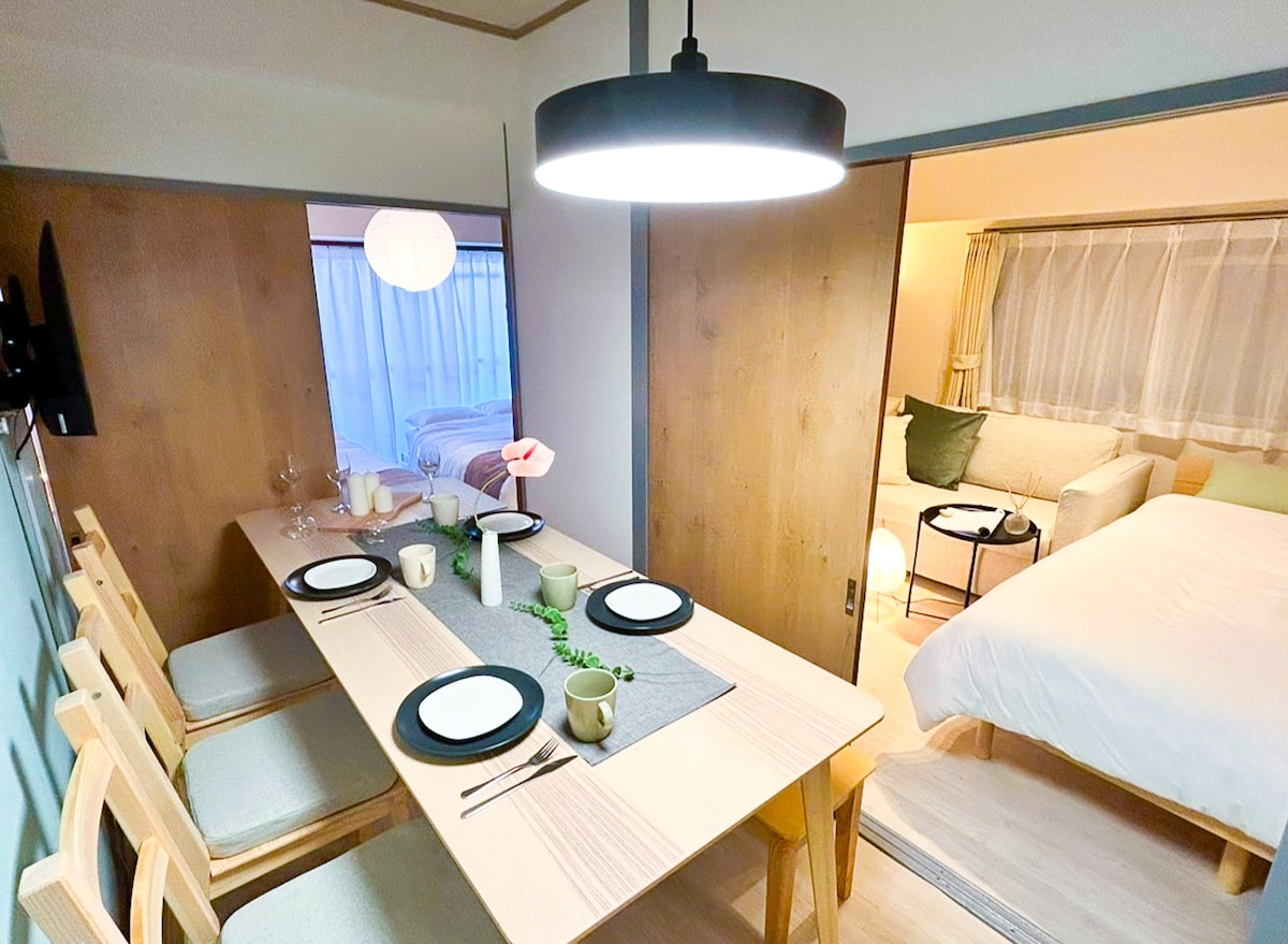 A dining area with a wooden table set for six is featured. Soft lighting from a pendant lamp illuminates the space. In the background, a cozy living area with a sofa and bedroom is visible, creating an inviting layout for shared moments.