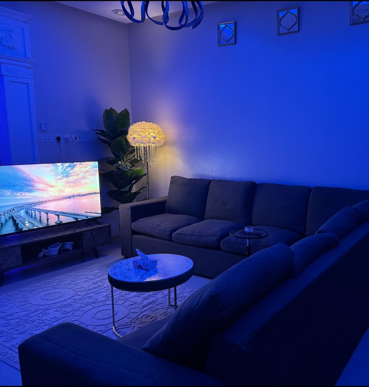 A modern living area is featured, illuminated by soft blue lighting. A large television displays scenic imagery, while a pair of gray sofas are positioned around a small round table. A decorative plant and a unique light fixture contribute to the overall ambiance.