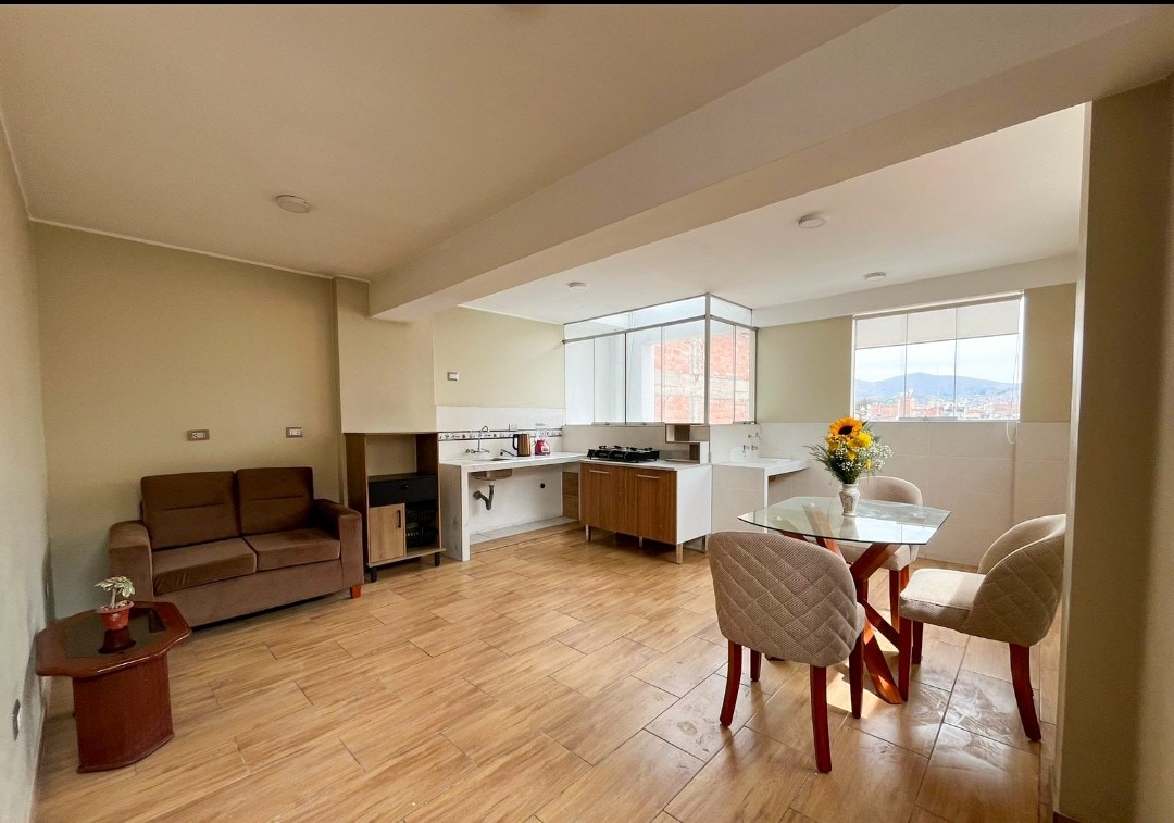 The open living space features a cozy sofa facing a dining area with a small table and four chairs. A well-equipped kitchen area is visible, with natural light streaming through large windows, offering a view of the surrounding area. A vase with flowers adds a decorative touch.
