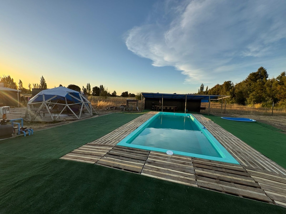 A well-maintained swimming pool is situated on a sunny lawn surrounded by grass. Wooden decking enhances the pool area, providing ample space for relaxation. A spacious geodesic dome structure is visible in the background, with clear skies and natural landscapes creating a calming atmosphere.