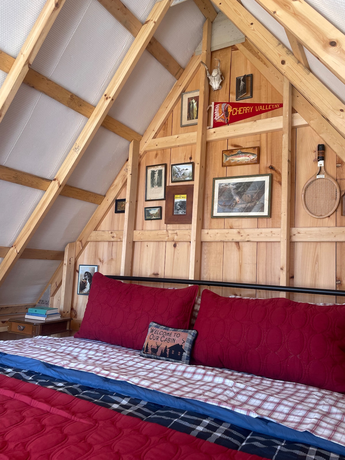 The sleeping area features a spacious bed with a deep red bedspread and decorative pillows. The A-frame walls are adorned with wooden paneling and a collection of framed pictures, alongside vintage items like a tennis racket. Natural light filters through the angled windows above.