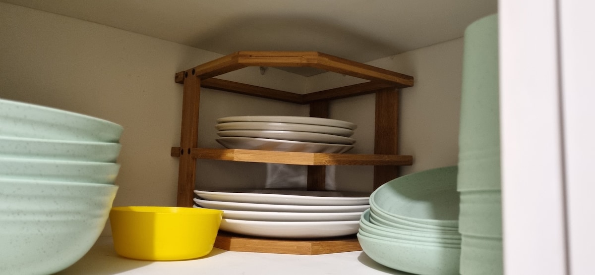 A well-organized kitchen shelf displays a variety of dishes. Stacked white plates rest atop a bamboo rack, while light green and a bright yellow bowl are positioned on the lower level, showcasing a functional and neat arrangement.