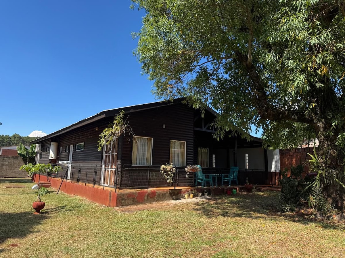 A spacious dark wood house is surrounded by a lush green lawn. Large windows allow natural light to fill the interior. A covered porch with bright blue seating is visible, providing a comfortable outdoor space for relaxation. Nearby trees offer shade and enhance the tranquil environment.