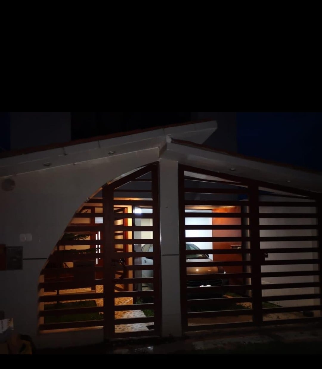 The entrance features a modern gate with horizontal wooden slats, allowing glimpses of the well-lit interior. A car is parked in the garage, framed by soft lighting that highlights the architecture and clean lines of the facade.