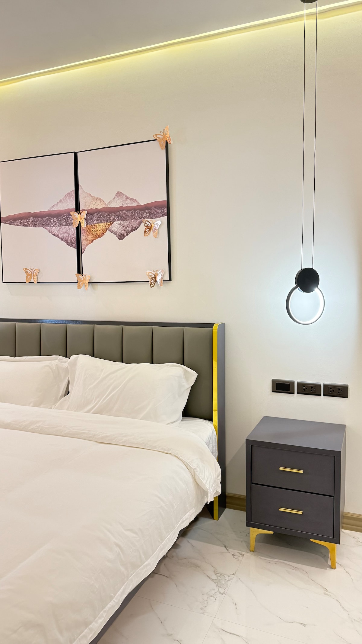 The bedroom features a stylish headboard with a sleek, gold accent. Above the bed, artwork displaying mountainous landscapes is adorned with decorative butterflies. A bedside table with two drawers stands next to the bed, and a modern pendant light provides gentle illumination.